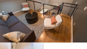 Un loft moderno con un sofá de color oscuro, una mesa redonda de centro y una silla blanca con cojín. Una escalera con barandilla conduce a un nivel superior.