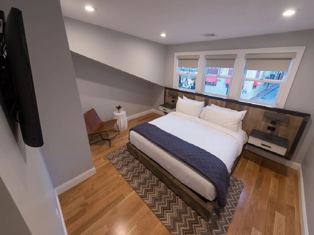 A bedroom with a large bed, a chair, a small table, and a window with a view of the street.