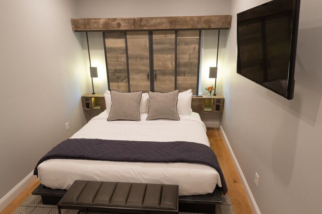 Ein modernes Hotelzimmer mit einem großen Bett, zwei Nachttischen mit Lampen, einem Flachbildfernseher und einer Bank am Fußende des Bettes.