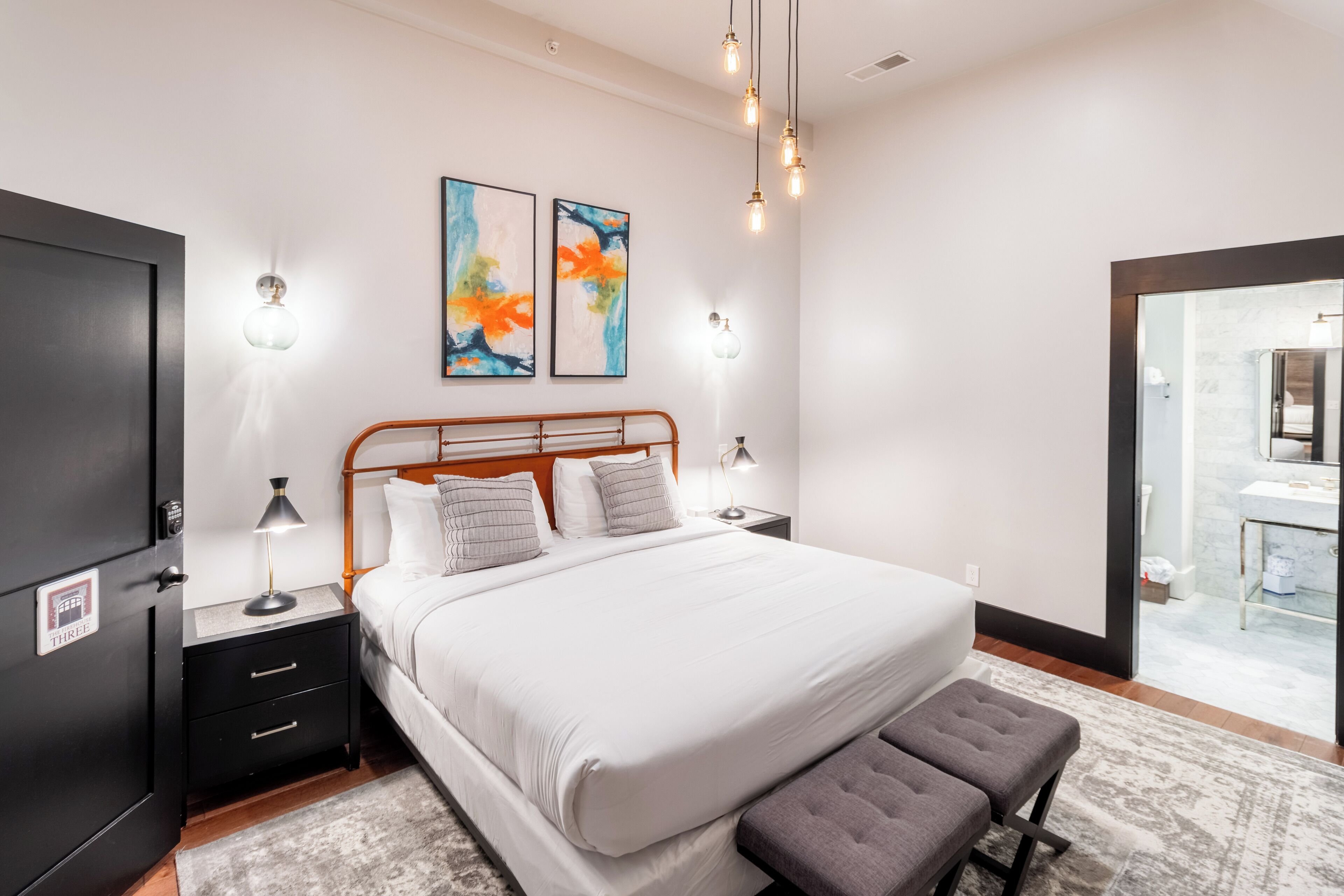 A modern bedroom with a large bed, bedside tables, a bench, artwork, and a bathroom visible through an open door.