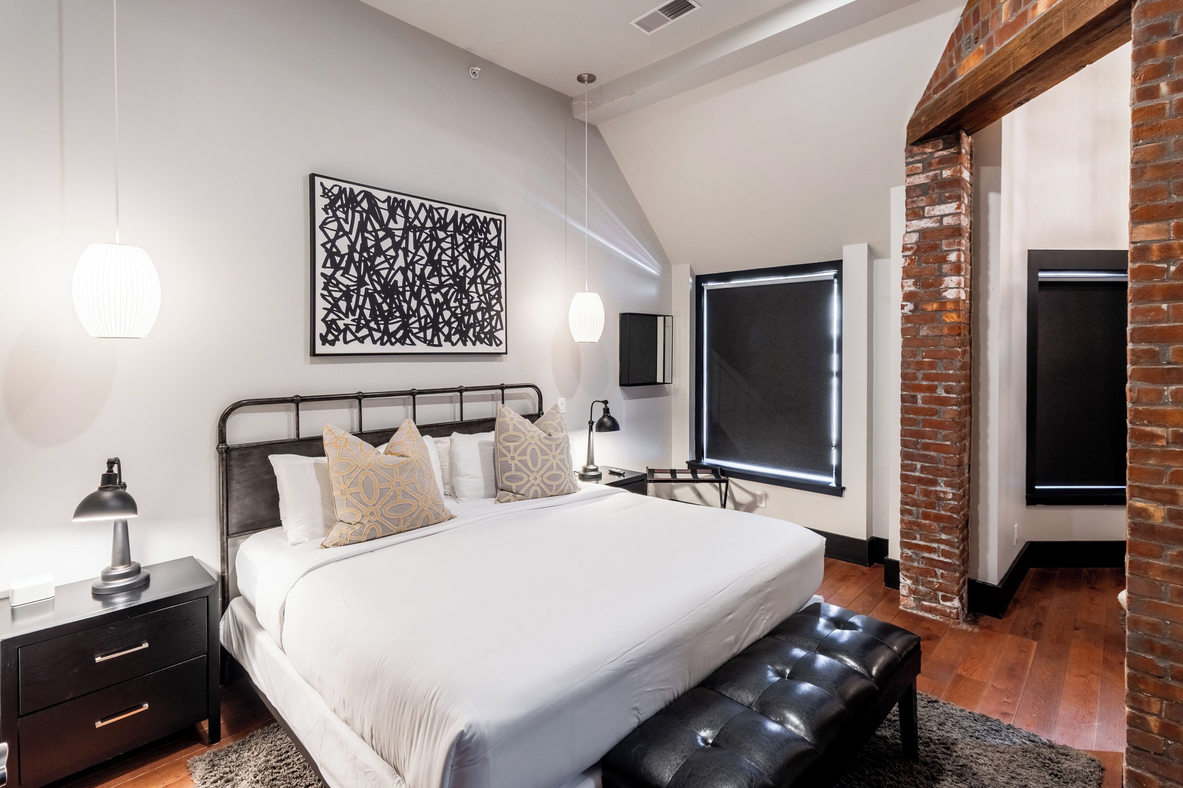 Une chambre moderne avec un grand lit, une table de chevet, un banc, un téléviseur fixé au mur et un mur d’accent en briques.