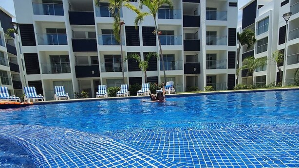 Ein Schwimmbecken mit Fliesenboden, umgeben von einem modernen Gebäude mit mehreren Balkonen und Liegestühlen.