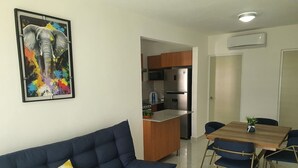 Ein modernes Wohnzimmer mit einem blauen Sofa, einem Holzesstisch mit Stühlen, einer Küchenzeile inklusive Kühlschrank und Mikrowelle sowie einem farbenfrohen Gemälde an der Wand.