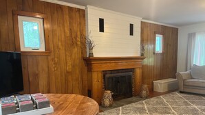 A living room with wooden paneling, a fireplace, a television, and a round table with magazines. - Fully Furnished Seneca Lake Lakefront Rental (Romulus)