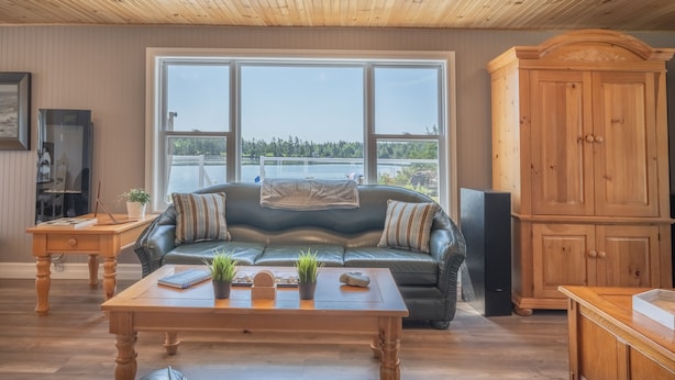 A cozy living room with a leather sofa, wooden coffee table, and a view of a lake through large windows. - Oceanfront Cottage on a Quiet Cove Just Outside Lunenburg (Lunenburg)