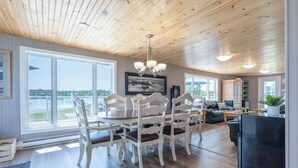 A spacious living area with a dining table and chairs, a kitchen area with a refrigerator, and large windows offering a view of the outdoors. - Oceanfront Cottage on a Quiet Cove Just Outside Lunenburg (Lunenburg)