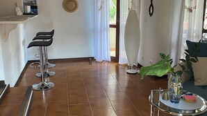 A spacious living area with a kitchen counter, bar stools, a large plant, and a glass table with a tray. - ♥️-lich Willkommen im la Dolce Vita -charming house with garden and terrace - (Narbolia, Loc. Is Arenas)