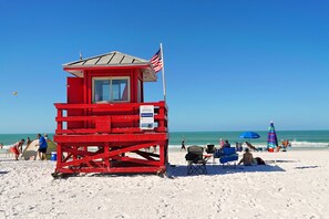 A red lifeguard tower with an American flag on a beach with people, a blue umbrella, and a tent.
