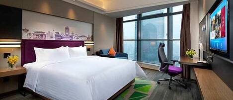 A modern hotel room with a large bed, a desk with a chair, a television, and a cityscape mural on the wall.