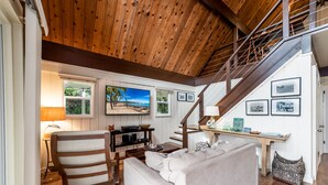 A spacious living room with a high wooden ceiling, a staircase, and a large window with a view of the outdoors. - Private Country Cottage | 2+ Bedroom on 1-acre | 5 mins to Kamuela town (Waimea)