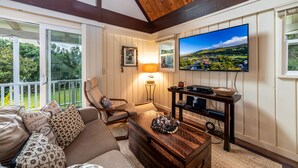 A cozy living room with a wooden ceiling, a large TV mounted on the wall, a comfortable sofa with patterned cushions, a coffee table, and a lamp. - Private Country Cottage | 2+ Bedroom on 1-acre | 5 mins to Kamuela town (Waimea)