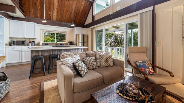 A spacious living area with a high ceiling, wooden beams, a large window, a sofa with patterned cushions, a chair, and a coffee table with snacks. - Private Country Cottage | 2+ Bedroom on 1-acre | 5 mins to Kamuela town (Waimea)