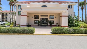 Entrada de un hotel con un corredor techado, palmeras y un vehículo estacionado afuera.