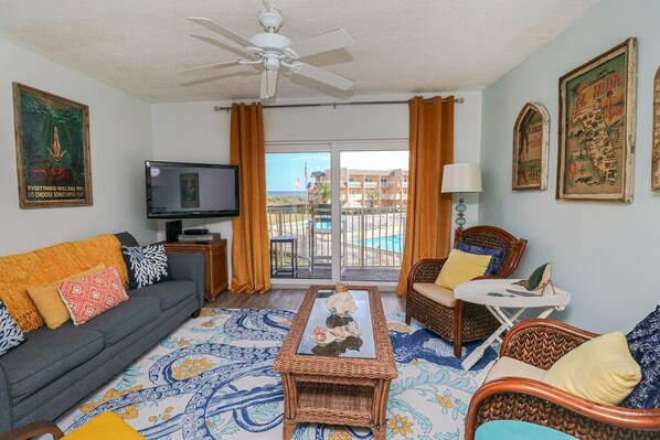 A living room with a sofa, armchairs, a coffee table, a television, and a balcony with a pool view. - Creston House 3E- Ocean View Two Bedroom Townhome (St. Augustine)