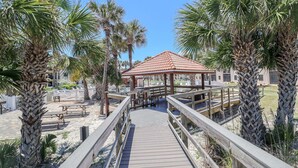 A wooden boardwalk leading to a gazebo with a tiled roof, surrounded by palm trees and a sandy area with picnic tables. - Ocean Villlage Club Condo G16 (St. Augustine)