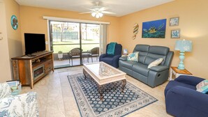 A living room with a television, sofa, coffee table, and a view through a sliding door to an outdoor area. - Ocean Villlage Club Condo G16 (St. Augustine)