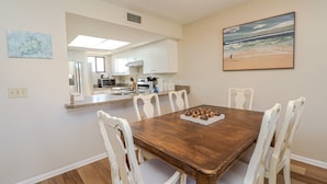 Uma área de refeições com mesa de madeira e cadeiras brancas, uma cozinha ao fundo e uma pintura com vista para a praia na parede.
