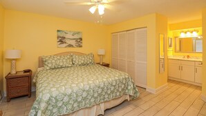 A bedroom with a bed, a nightstand, a ceiling fan, a painting, a closet, and a bathroom with a sink and mirror.