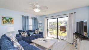 A living room with a sofa, coffee table, and TV. There is a balcony with a view of buildings and palm trees. - Ocean Village Club Condo R28 (St. Augustine)