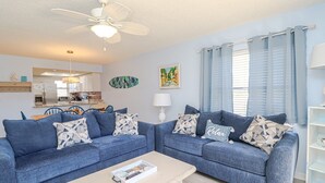 A living room with two blue sofas, a coffee table, a ceiling fan, and a kitchen in the background. - Ocean Village Club Condo R28 (St. Augustine)
