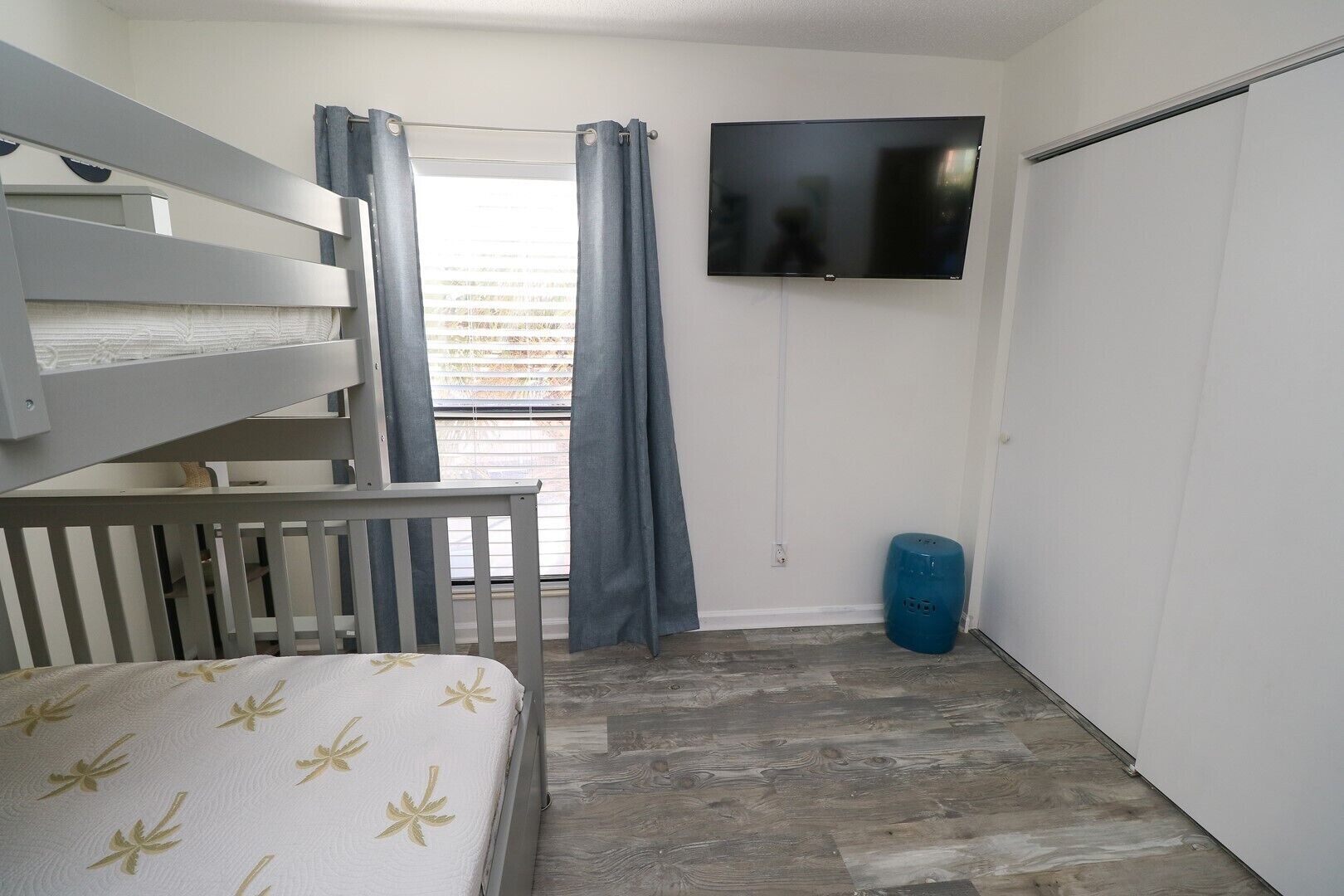 Ein Zimmer mit einem Etagenbett, einem an der Wand montierten Fernseher, einem Fenster mit Vorhängen und einem blauen Mülleimer.