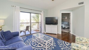 Ein Wohnzimmer mit blauem Teppichmuster, einer Couch, einem Fernseher auf einer weißen Kommode und einem Balkon mit einem Sessel.
