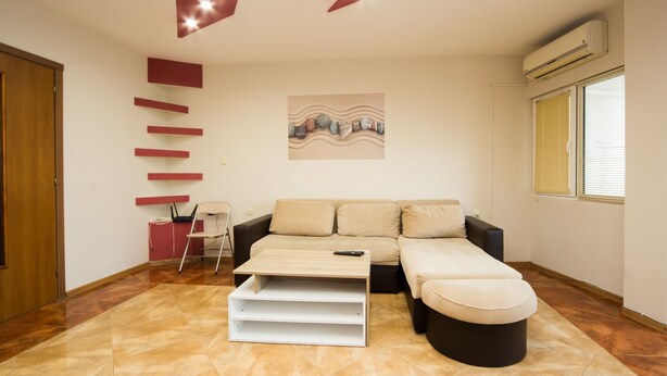Een moderne woonkamer met een bank, een poef en een witte salontafel. Er hangt een schilderij aan de muur, staat een fauteuil en een rode kast.