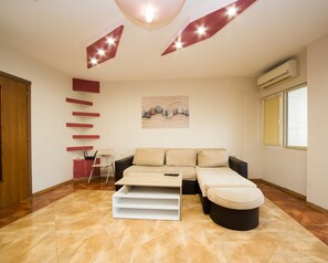 A modern living room with a sofa, ottoman, and a white coffee table. There is a painting on the wall, a chair, and a red shelf unit. - Aleksapart (Burgas)