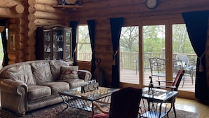 A log cabin interior with a leather sofa, dining table, chairs, and a large window overlooking a deck.