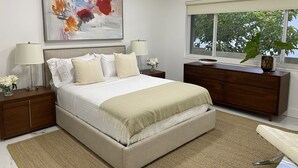 A modern bedroom with a large bed, two bedside tables with lamps, a dresser, a large window with a view of greenery, and a painting on the wall. - Waterfront condo overlooking Palm Beach (West Palm Beach)