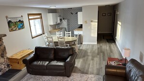Uma sala de estar com um sofá de couro marrom, um tapete estampado e um espaço de jantar com mesa e cadeiras de madeira.
