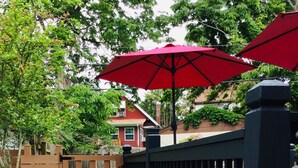 Un sentier bordé de pierres plates qui mène à une zone délimitée par une clôture en bois et agrémentée d’un parasol rouge.