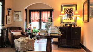 Un salon chaleureux avec un fauteuil en cuir, un petit pouf, une table ronde ornée de livres, un vase et une lampe. On y trouve également une bibliothèque avec des objets décoratifs et un tableau encadré accroché au mur.