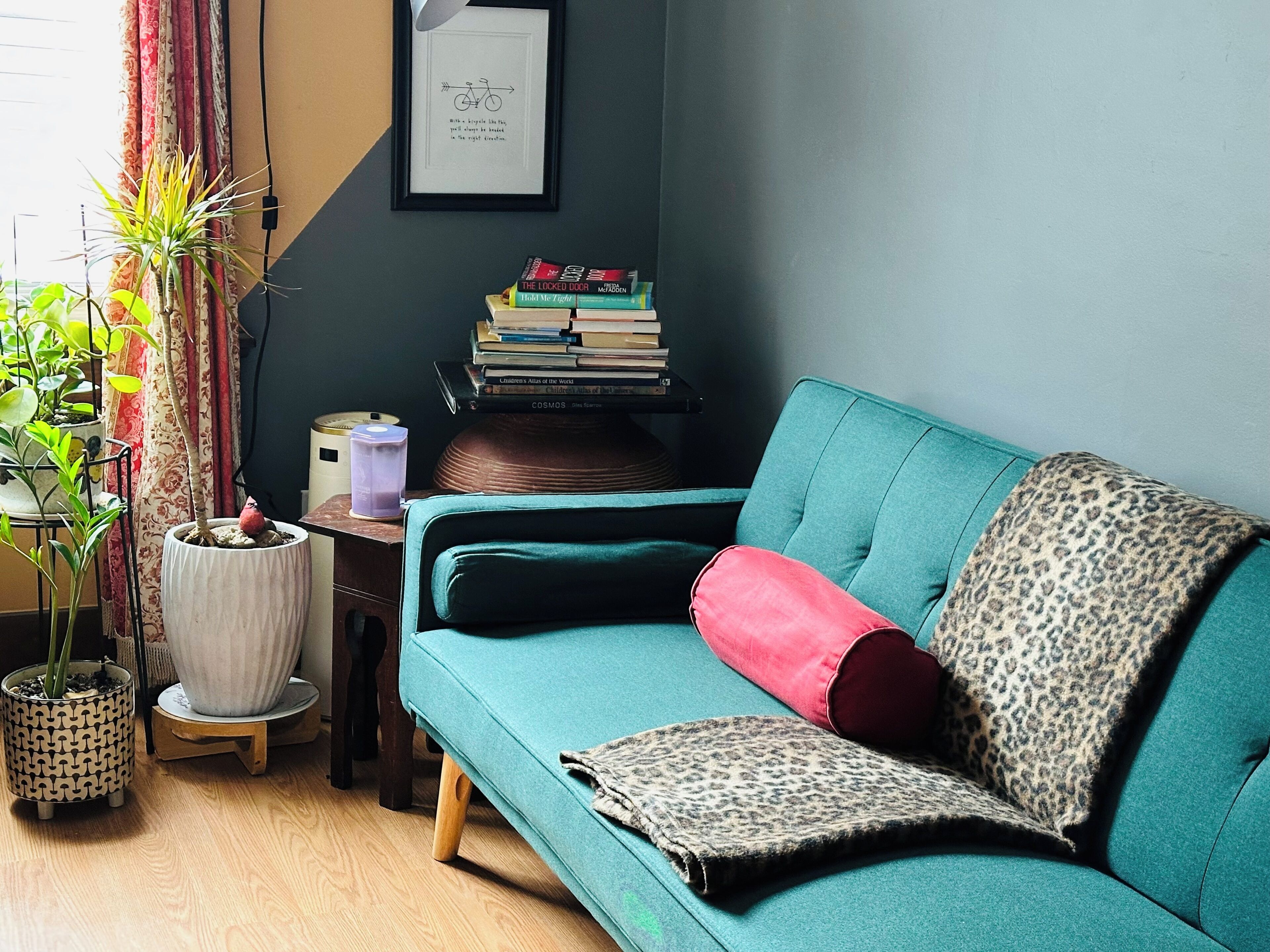 Ein smaragdgrünes Sofa mit Leopardenmuster und roten Kissen, ein Stapel Bücher sowie ein gerahmter Spruch an der Wand.