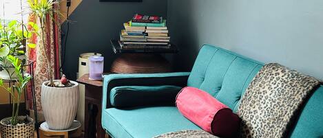 Ein smaragdgrünes Sofa mit Leopardenmuster und roten Kissen, ein Stapel Bücher sowie ein gerahmter Spruch an der Wand.