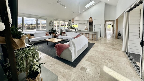 A spacious living area with a large window, a ceiling fan, and a kitchen area with a central island. - Coastal Retreat - POOL,  SPA & FIRE. Nature boardwalk, creek & inlet. 5 bedroom. (Inverloch)