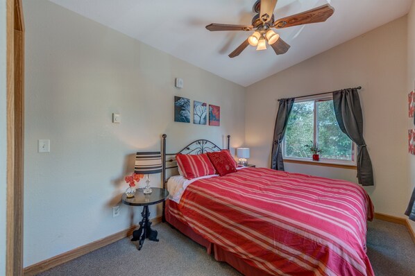 A bedroom with a bed, a nightstand, a ceiling fan, a window with curtains, and artwork on the wall.
