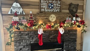 Uma lareira com moldura de pedra, decorada com guirlandas natalinas e meias de Natal. Acima da lareira, existem placas com mensagens festivas e uma decoração de urso.