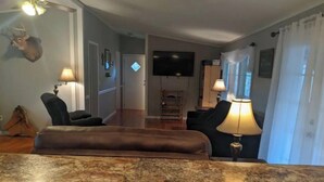 Uma sala de estar com um sofá de couro marrom, duas poltronas pretas, uma TV fixada na parede e uma lareira.