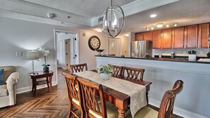 A dining area with a wooden table and chairs, a chandelier, and a kitchen in the background. - 5-Star Luxury, Fully Updated OCEANFRONT 2 Bed/ 2 Bath FALL STAYS 50% OFF RESORT! (Myrtle Beach)