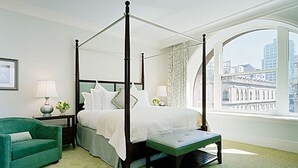 A four-poster bed with a bench, a green armchair, and a view of a cityscape through large windows.