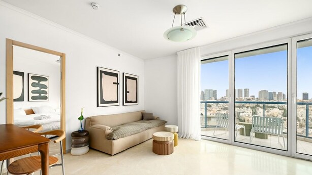 A modern living room with a sofa, a dining table, and chairs. There are framed pictures on the wall and a large window with a city view.