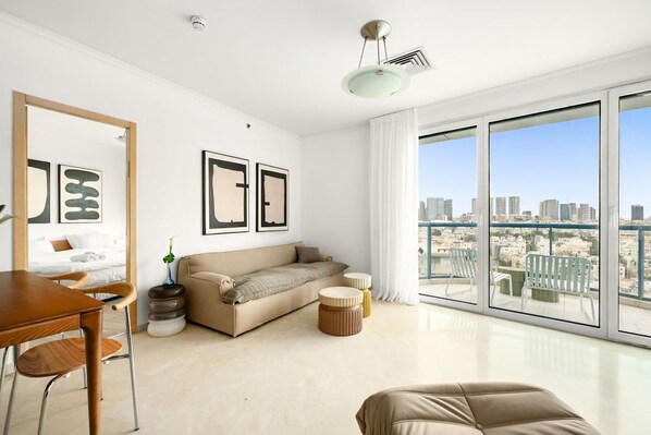 A modern living room with a sofa, a dining table, and chairs. There are framed pictures on the wall and a large window with a city view.