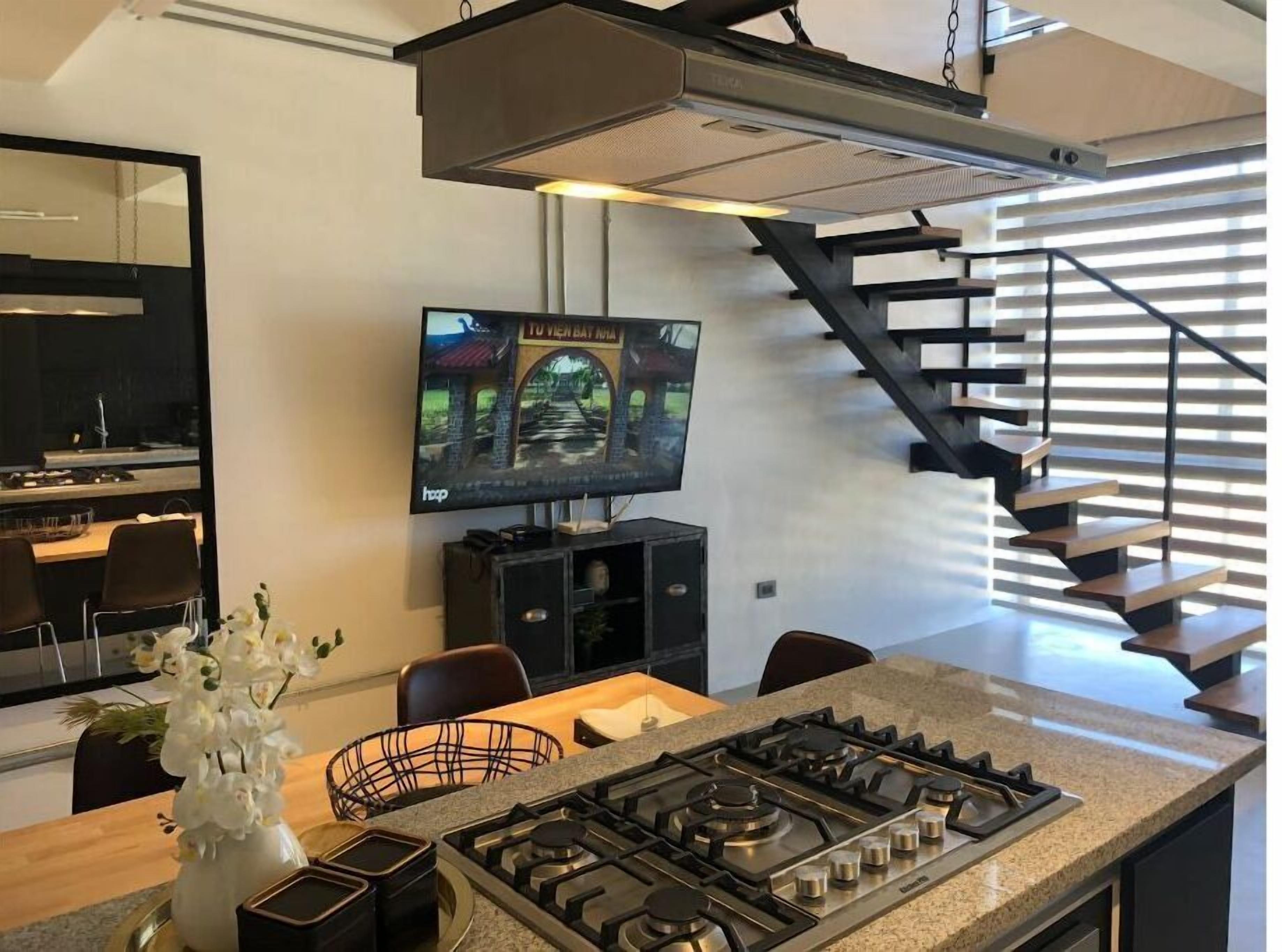 Una cocina moderna con estufa a gas, un comedor con sillas, un televisor montado en la pared y una escalera con barandilla de metal.