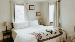 A neatly made bed with a patterned throw, two bedside lamps, and a chair in a simple, well-lit room with curtains and framed pictures on the walls.