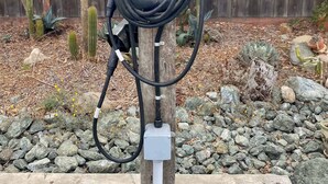 Una estación de carga para vehículos eléctricos al costado de la carretera, con una cerca de madera y cactus al fondo.