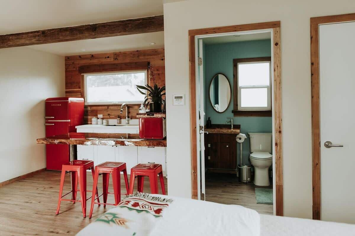 Una habitación compacta con cama, kitchenette con electrodomésticos rojos, techo de madera y baño a la vista a través de una puerta abierta.
