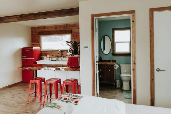 Una habitación compacta con cama, kitchenette con electrodomésticos rojos, techo de madera y baño a la vista a través de una puerta abierta.