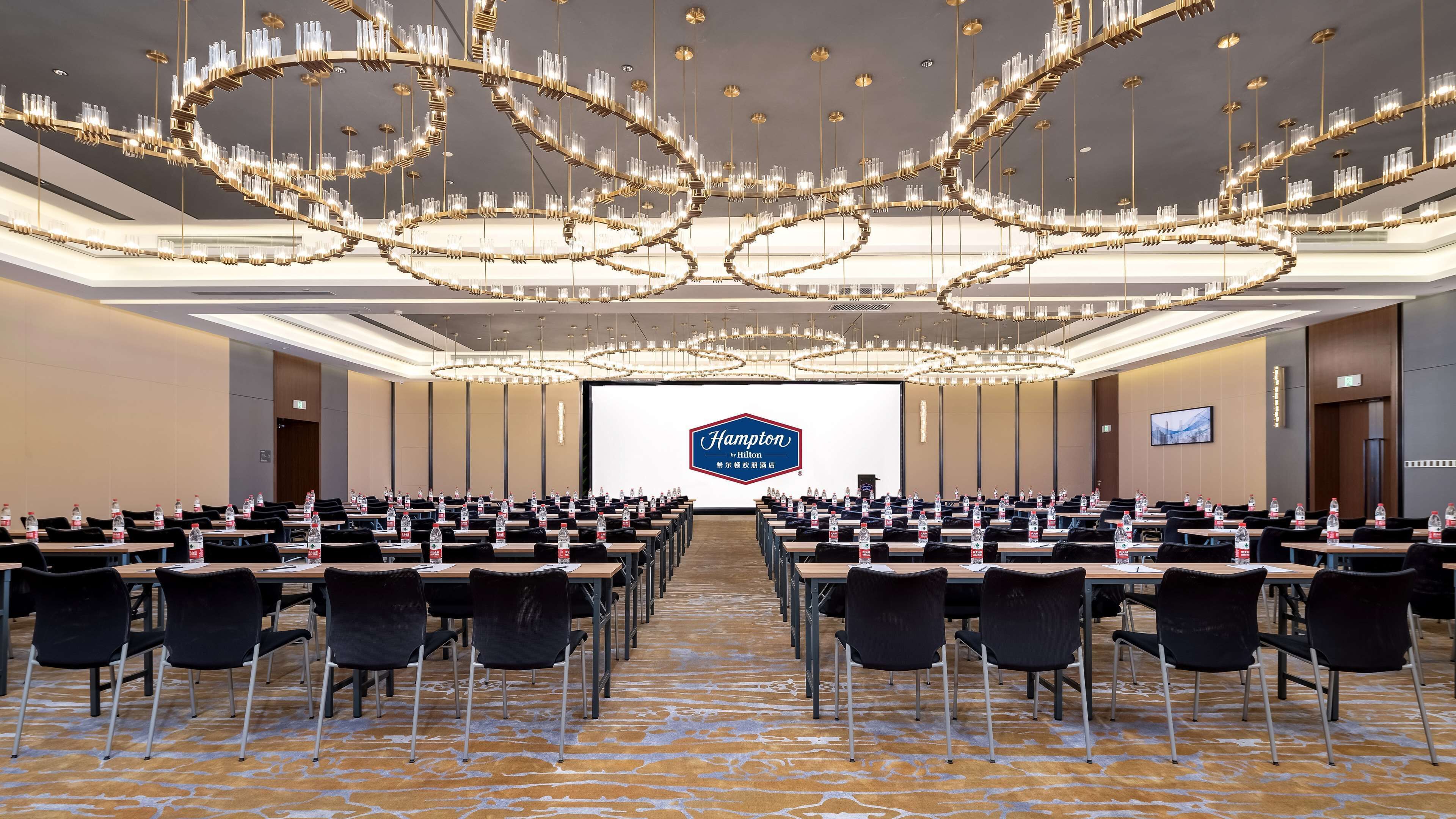 A conference room with rows of tables and chairs, a large screen displaying the Hampton by Hilton logo, and a modern chandelier.