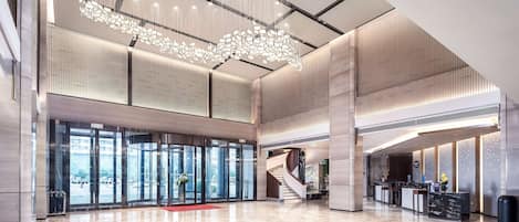 A spacious lobby with a modern chandelier, a reception desk, and a staircase leading to upper floors.